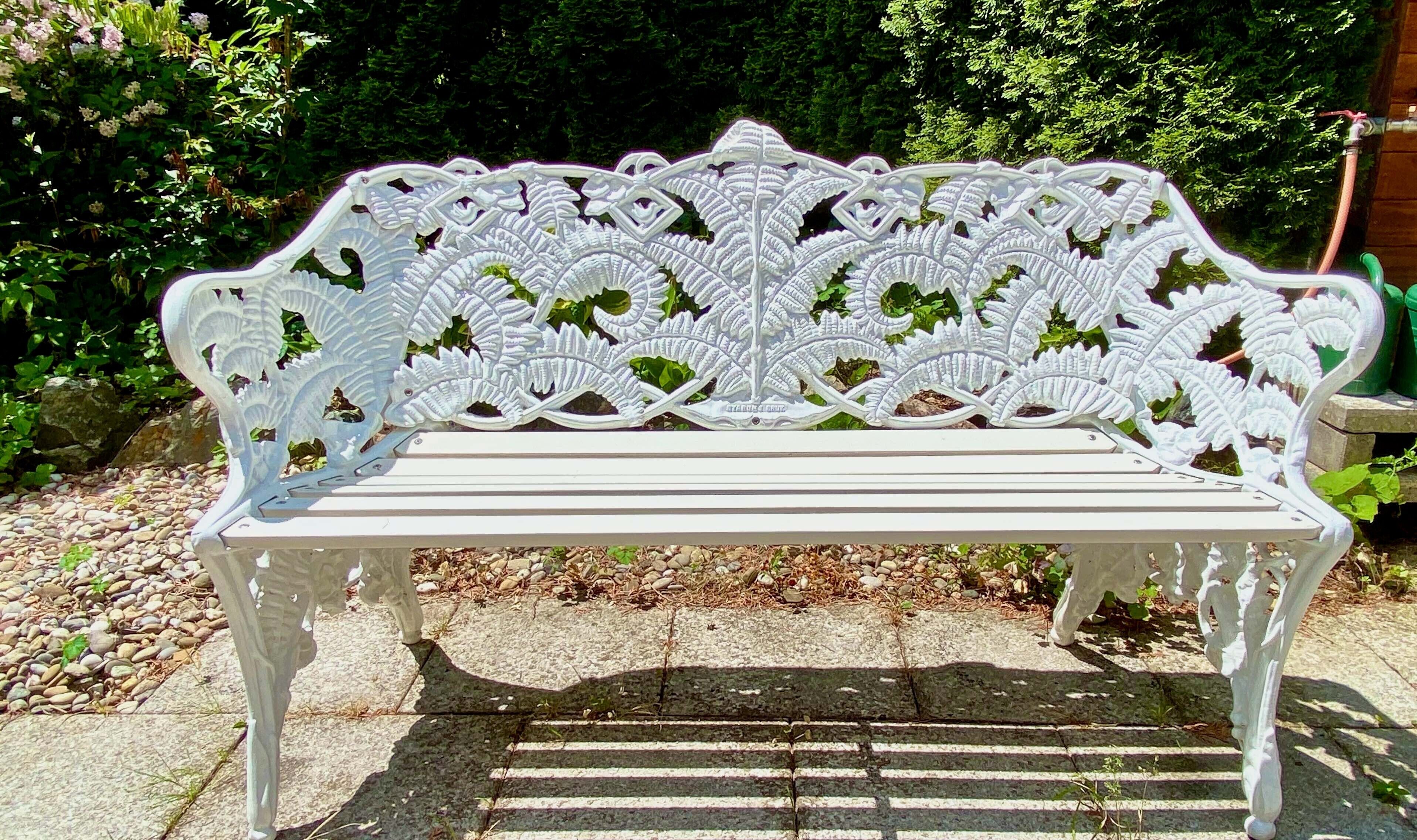 A white bench with intricate leaf patterns, set on a stone pathway in a garden with greenery and a hose in the background.