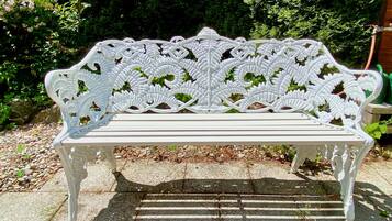 A white bench with intricate leaf patterns, set on a stone pathway in a garden with greenery and a hose in the background.