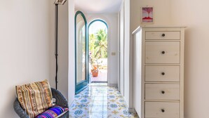 A hallway with a patterned floor, a wicker chair with a striped cushion, a tall floor lamp, and a white cabinet with drawers. - Capri Turquoise by Napoliapartments (Capri)