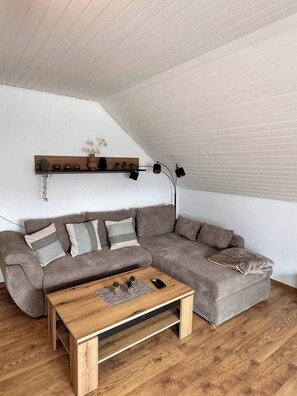 A modern living room with a sectional sofa, a wooden coffee table, and a wall-mounted shelf with decorative items. - Riding & recreation in the Weserbergland (Salzhemmendorf)