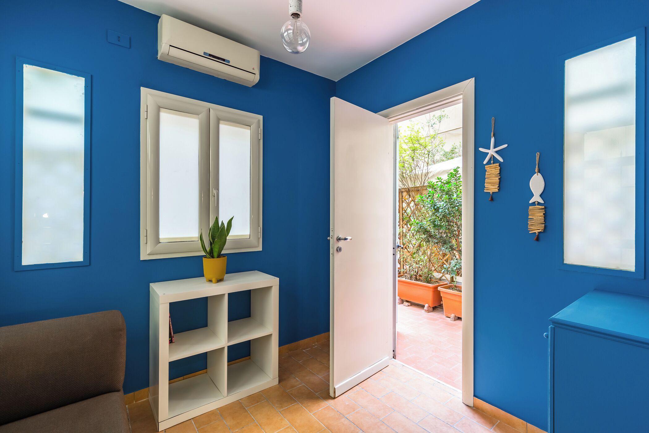 Une pièce aux murs bleus, avec une porte blanche, une étagère blanche ornée d’une plante et une fenêtre avec des stores.