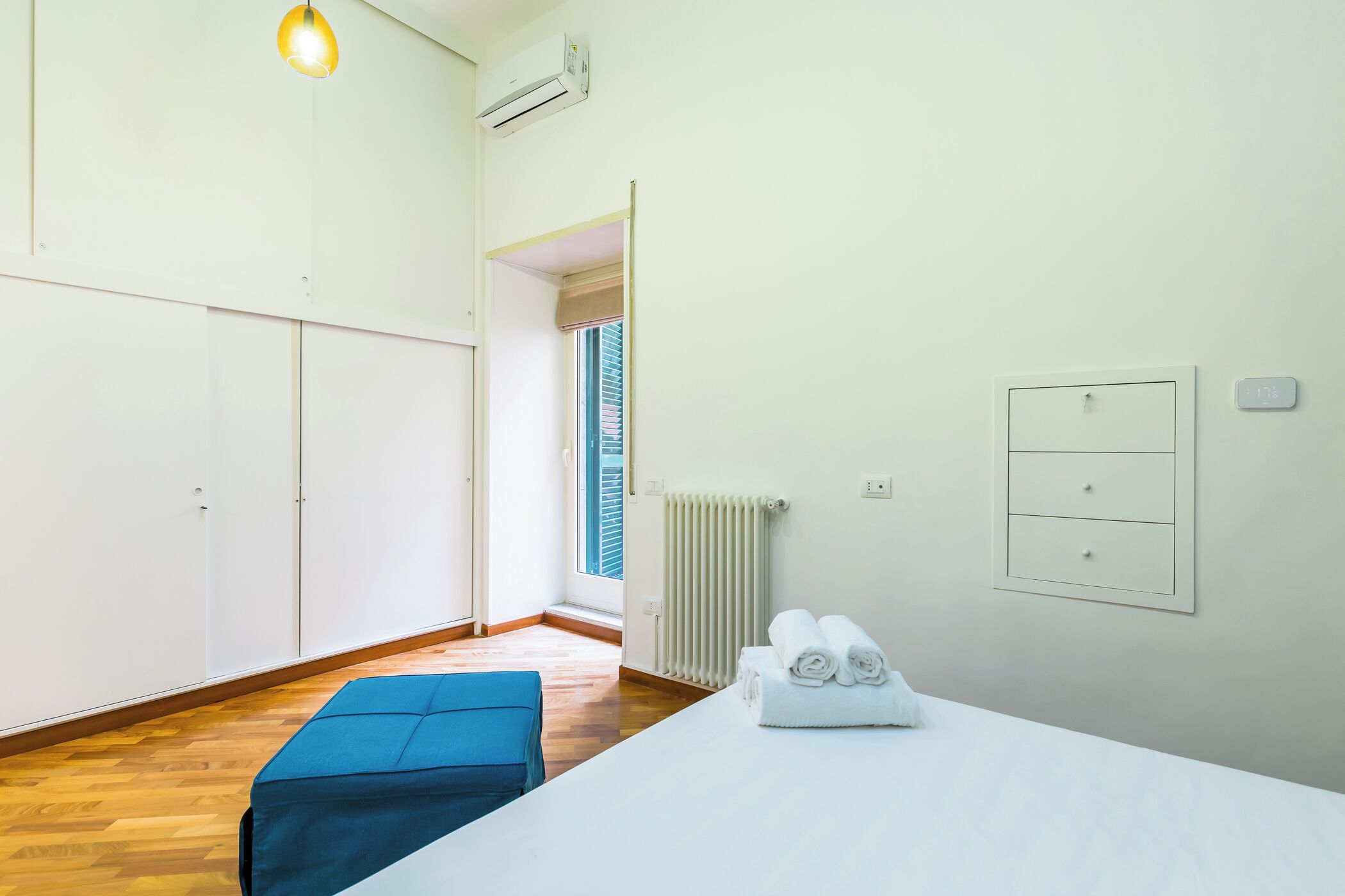 Un dormitorio moderno con piso de madera, una cama con sábanas blancas, un banco azul, un radiador y una ventana con persianas.