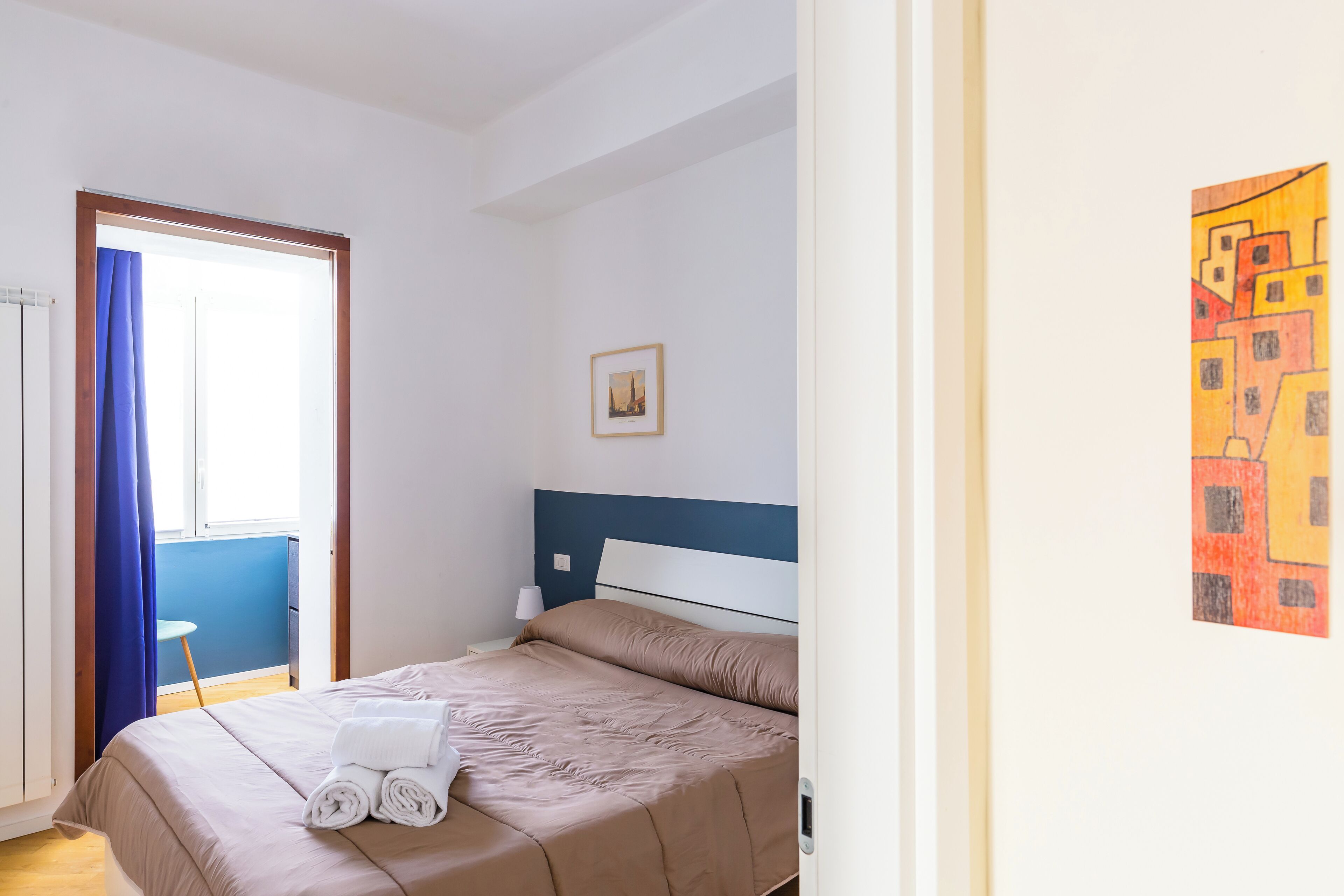 A neatly made bed with a blue headboard, a small bedside table with a lamp, and a framed picture on the wall.