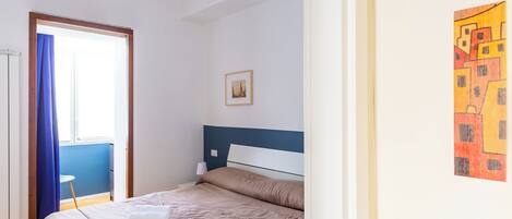 A neatly made bed with a blue headboard, a small bedside table with a lamp, and a framed picture on the wall.