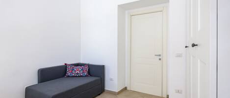 A small, dark-colored sofa with a colorful pillow against a white wall, a white door with a handle, and a tiled floor.
