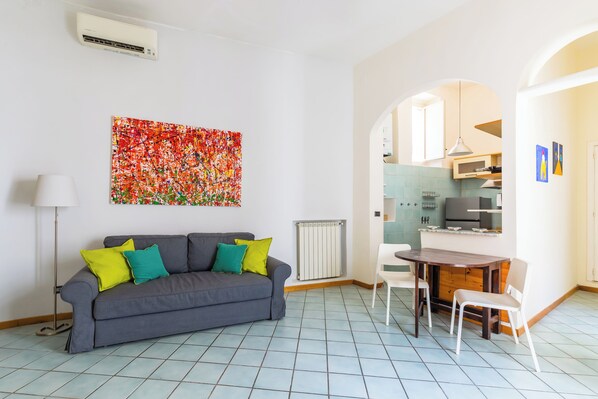 A living room with a grey sofa, a dining table with chairs, a radiator, and a kitchen visible through an archway. - Chiaia Relax on Pedastrian Area (Naples)