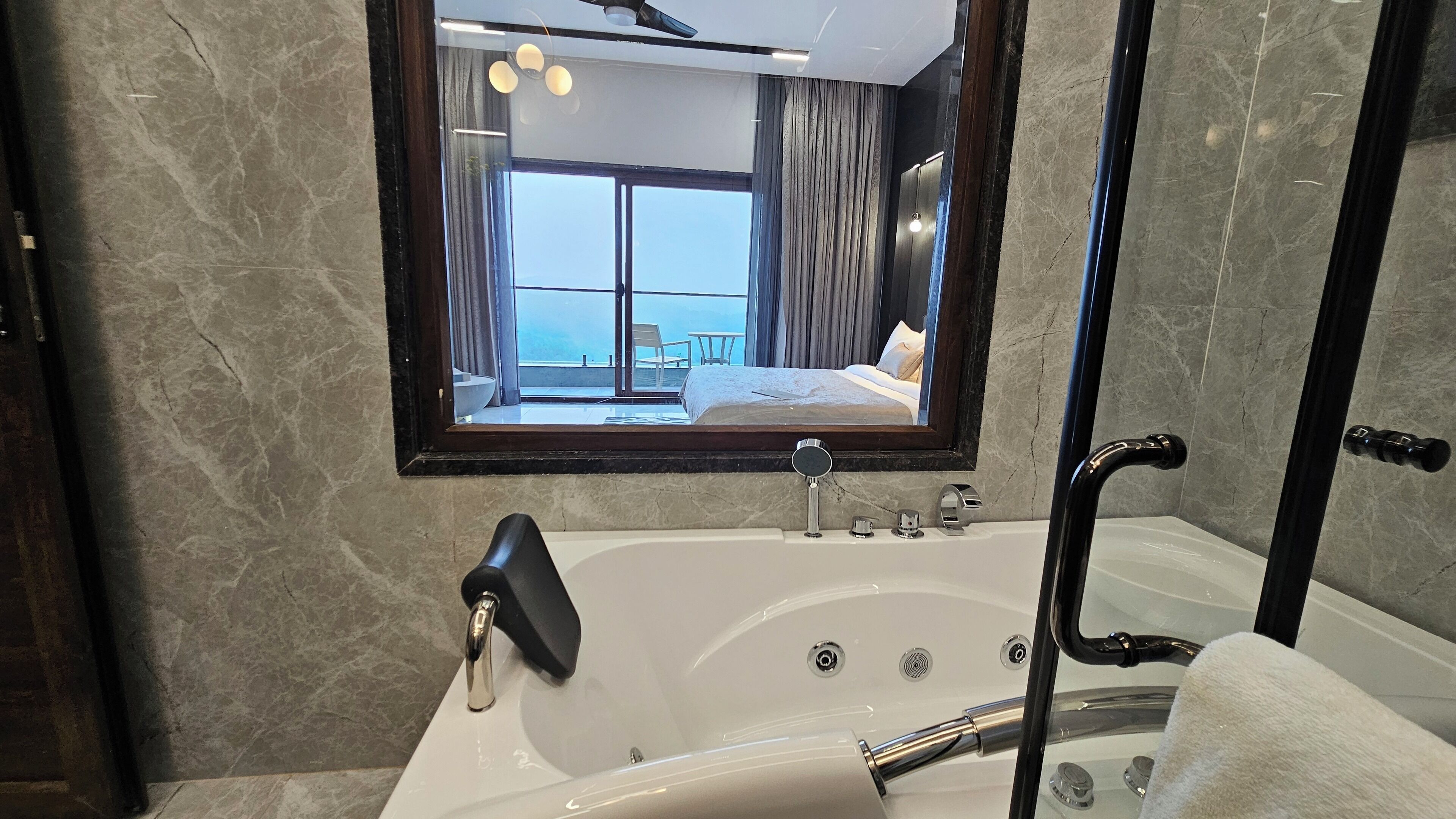 A modern bathroom with a large bathtub, a mirror, and a view of a balcony with a bed.