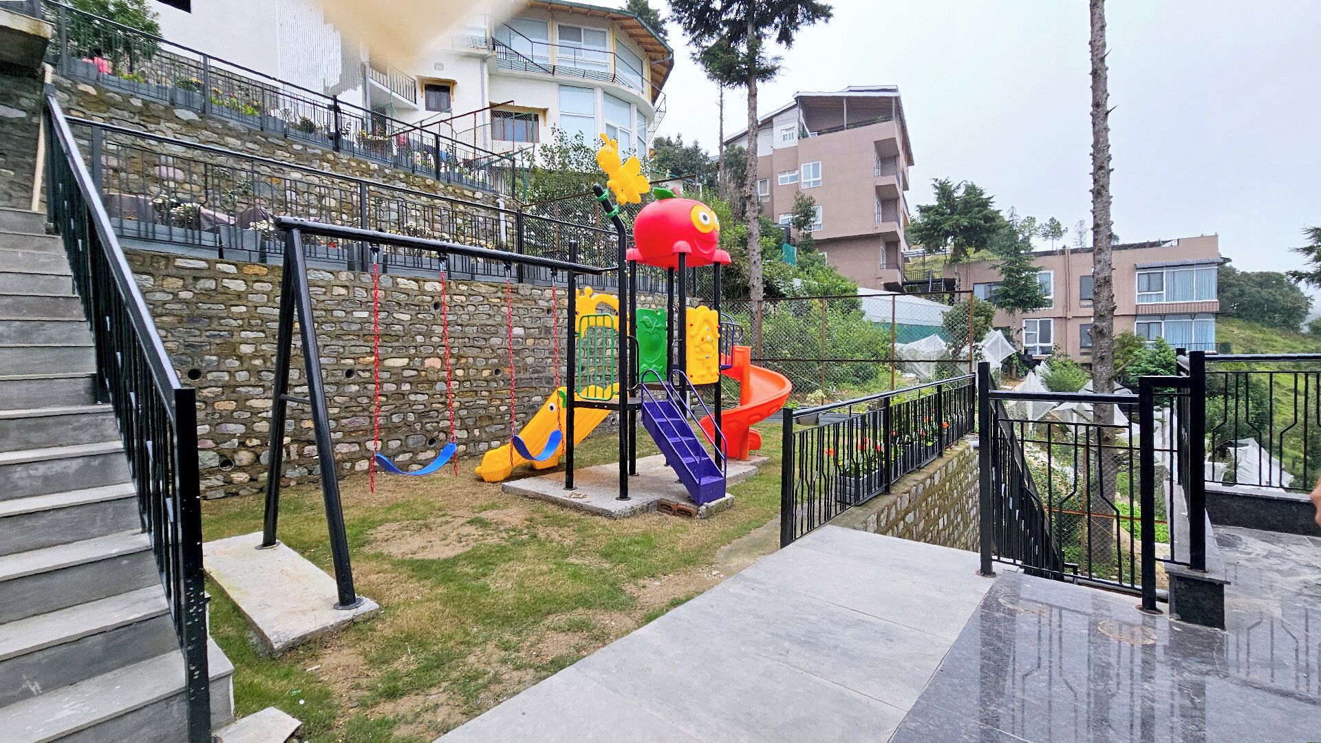 A playground with a slide and climbing structures, surrounded by stone walls and residential buildings.