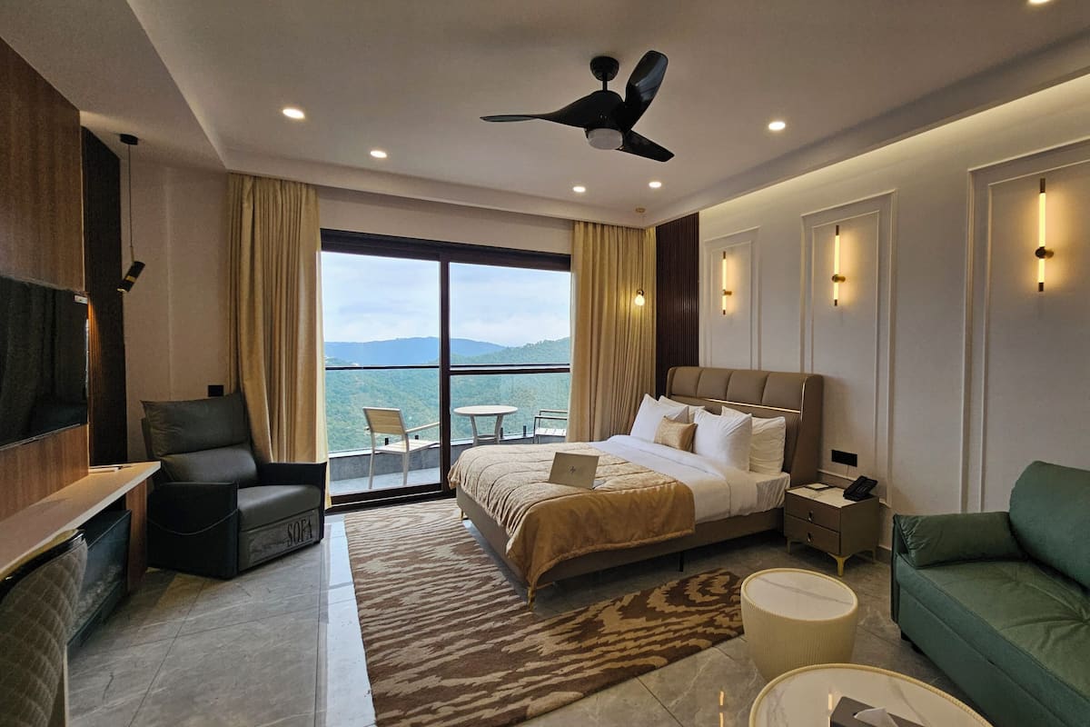 A hotel room with a bed, a sofa, a desk, a chair, a balcony with a view, and a ceiling fan.