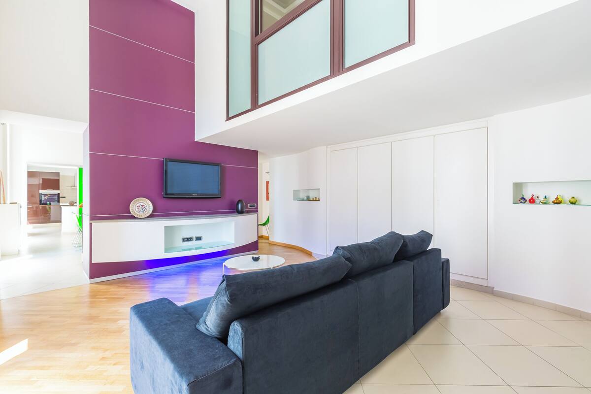 A modern living room with a dark grey sofa, a flat-screen TV mounted on a purple accent wall, and a small white coffee table.