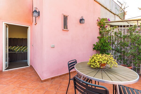 Ein rosafarbenes Gebäude mit einer Terrasse, auf der ein runder Tisch mit Stühlen, ein Blumentopf und ein kleines Fenster zu sehen sind.