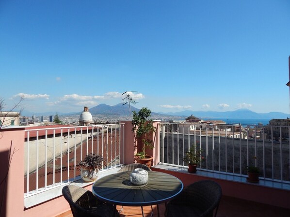 Terraço no telhado com uma mesa redonda e cadeiras, plantas em vasos e vista para a cidade e montanhas ao longe.