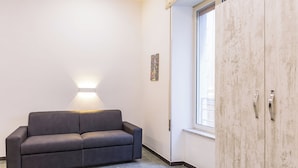 Una habitación moderna con un sofá gris oscuro, un armario de madera, una lámpara de pared y una ventana.