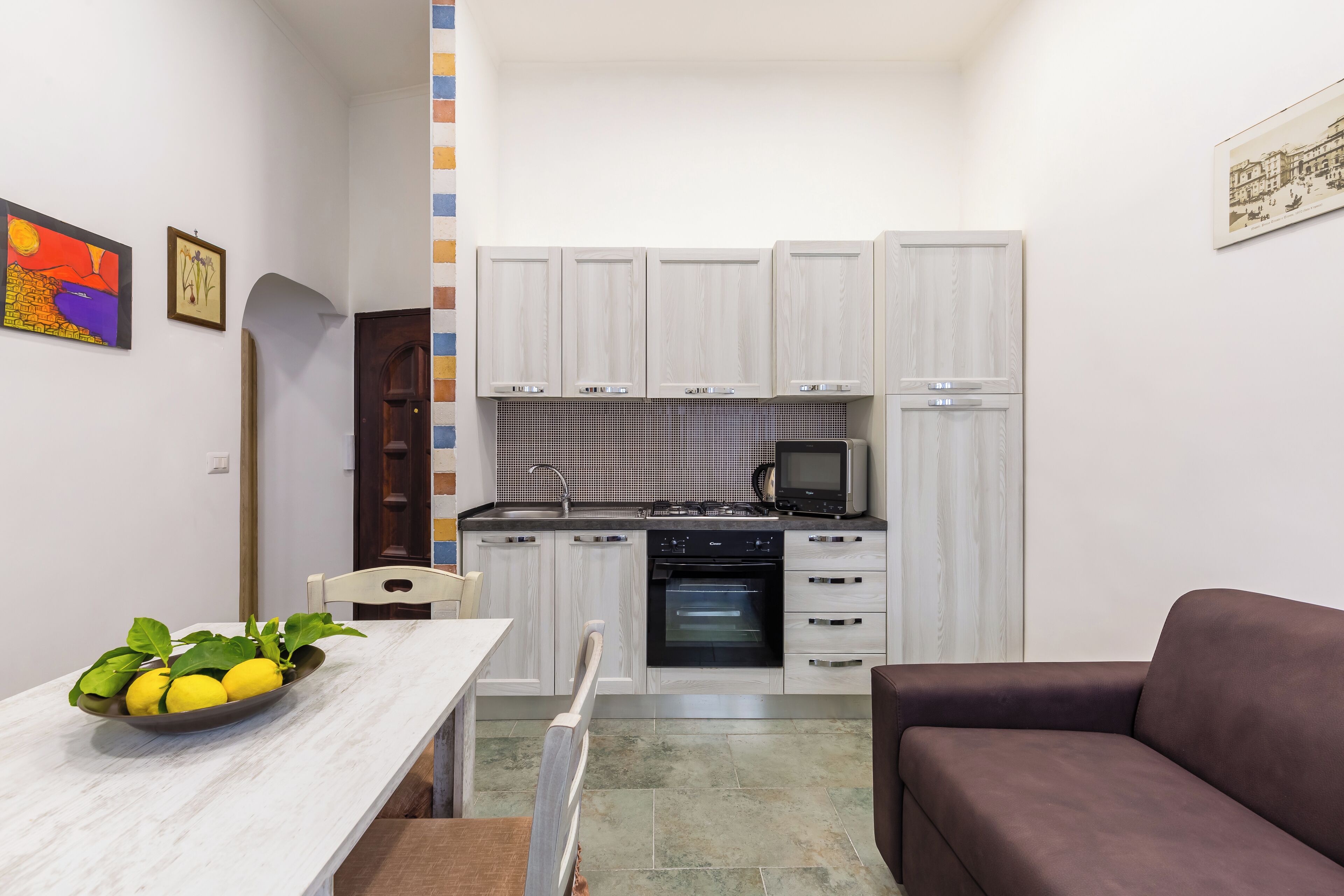 A compact kitchen with white cabinets, a microwave, and an oven. There is a dining table with a bowl of lemons and a chair.