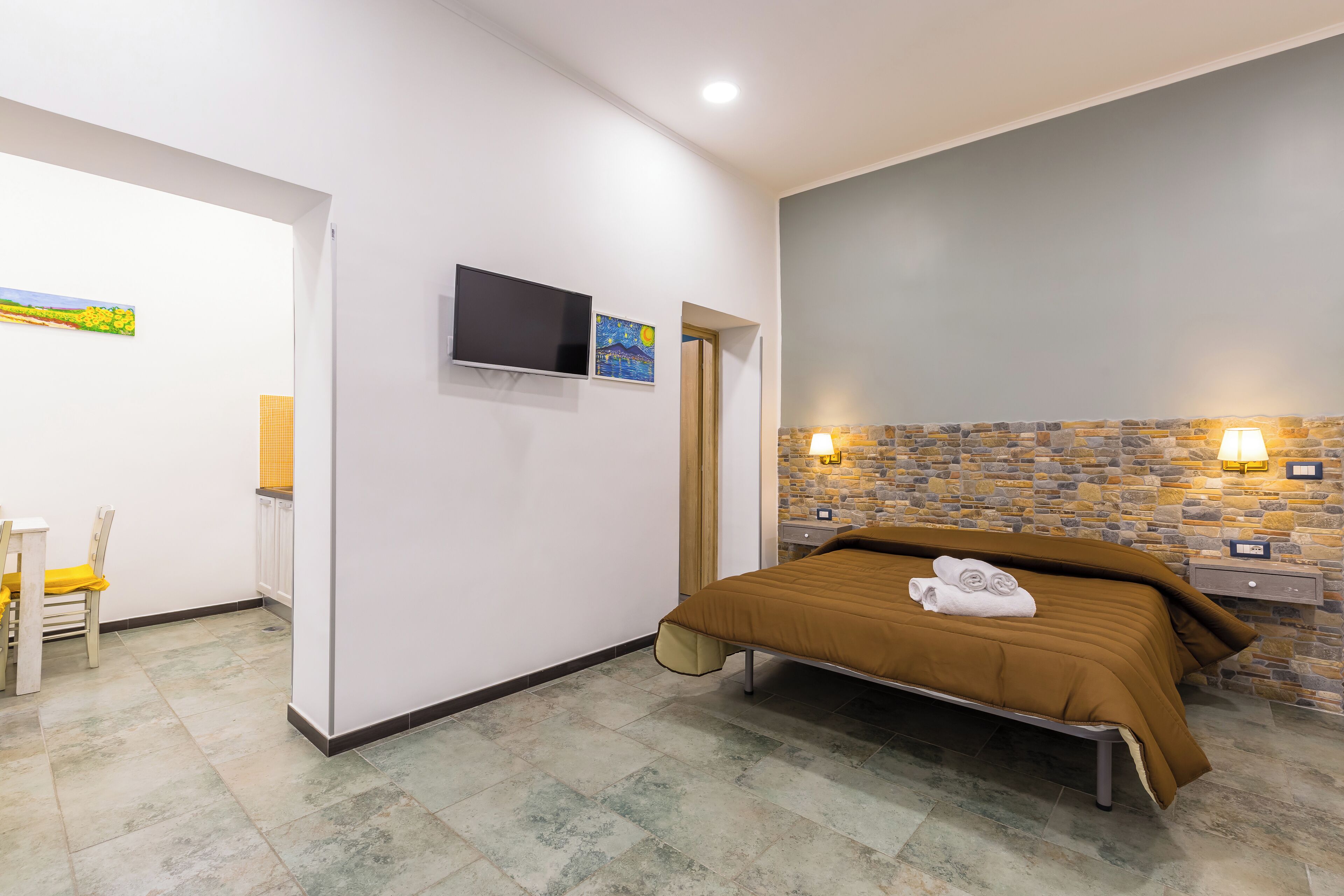 Una camera da letto con un letto, una TV, un quadro, un muro in pietra e un tavolino con una lampada.