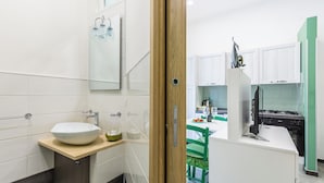 Un baño moderno con un lavabo blanco grande, un espejo y una puerta de madera que da a una zona de cocina con estufa y sillas verdes.
