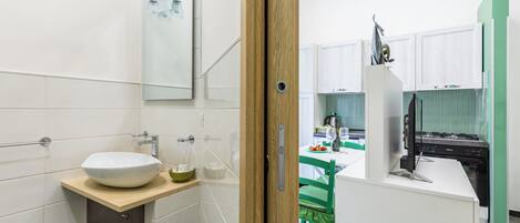 A modern bathroom with a large white sink, a mirror, and a wooden door leading to a kitchen area with a stove and green chairs.