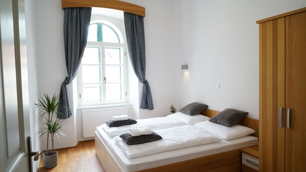 Ein Schlafzimmer mit einem hölzernen Bett, grauen Vorhängen, einem Fenster mit weißem Rahmen, einer kleinen Pflanze und einem Holzschrank.