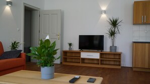 Ein modernes Wohnzimmer mit Flachbildfernseher auf einem Holzboden, einem Esstisch mit Fernbedienung und Topfpflanzen.