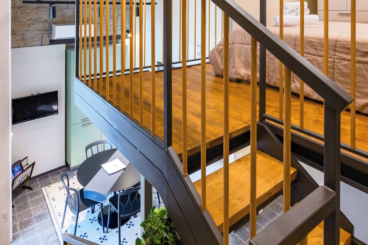 Een moderne loft met zichtbare houten balken, een trap, een eetgedeelte en een slaapkamer met een bed en nachtkastje.