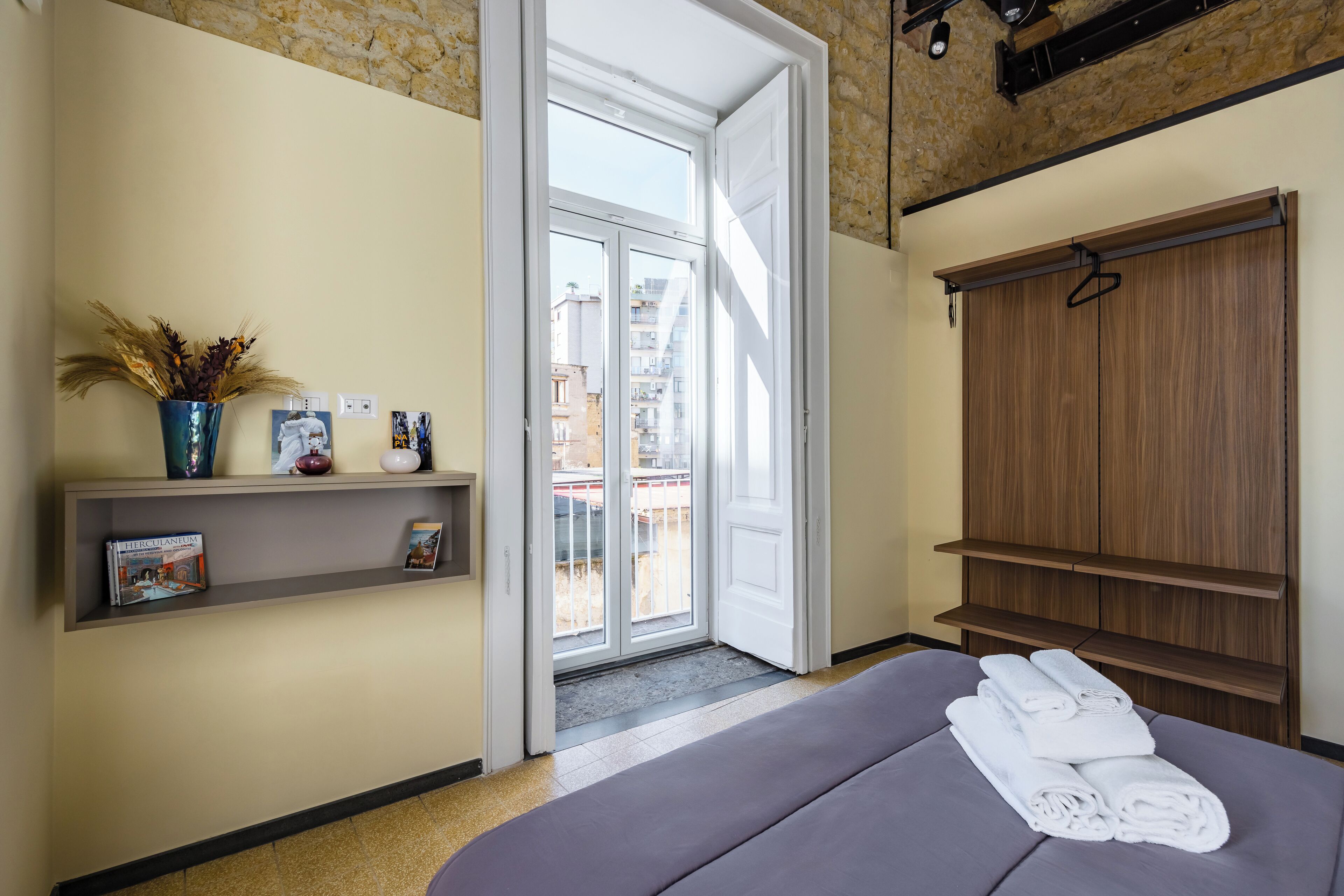 Habitación de hotel con cama, una estantería con libros, un florero con plantas secas y vistas al balcón exterior.