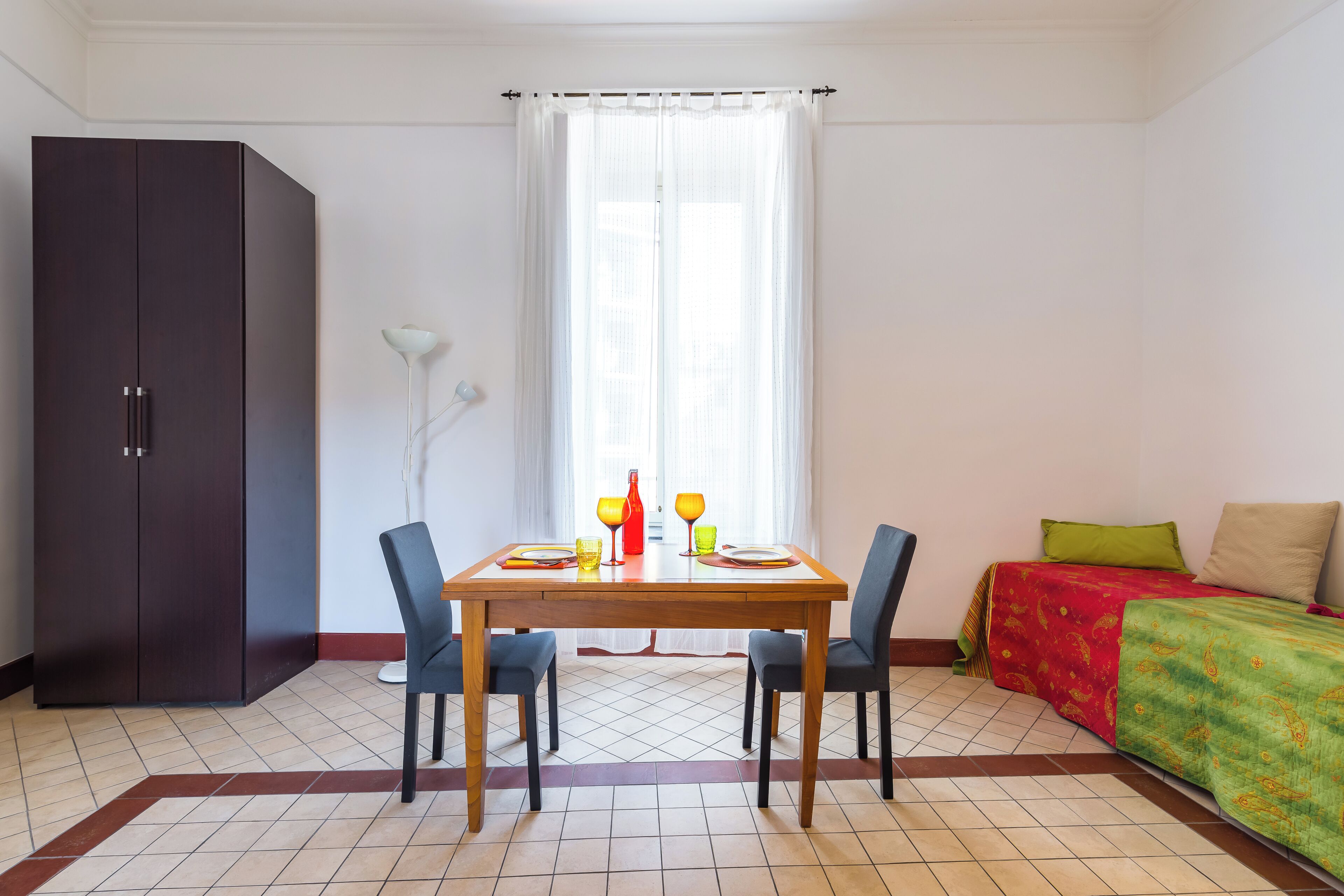 Una stanza con un tavolo da pranzo apparecchiato per due persone, un armadio, un letto con copriletto colorato e una finestra con tende leggere.