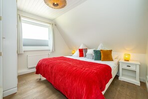 A bedroom with a sloped ceiling, a large window, a bed with a red bedspread, multiple pillows, and bedside lamps.