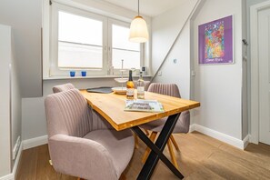 Un coin repas comprenant une table en bois, deux chaises rembourrées, un luminaire suspendu et une fenêtre avec des stores.