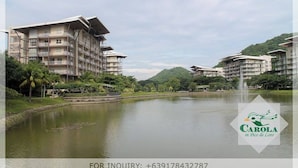 A modern building complex with multiple balconies overlooking a calm lake, surrounded by lush greenery and hills.