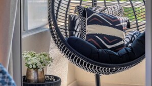 A hanging chair with a cushion, a small table with a vase of flowers, and a view of the sky and buildings in the background. - Voula Urban Escape (Voula)