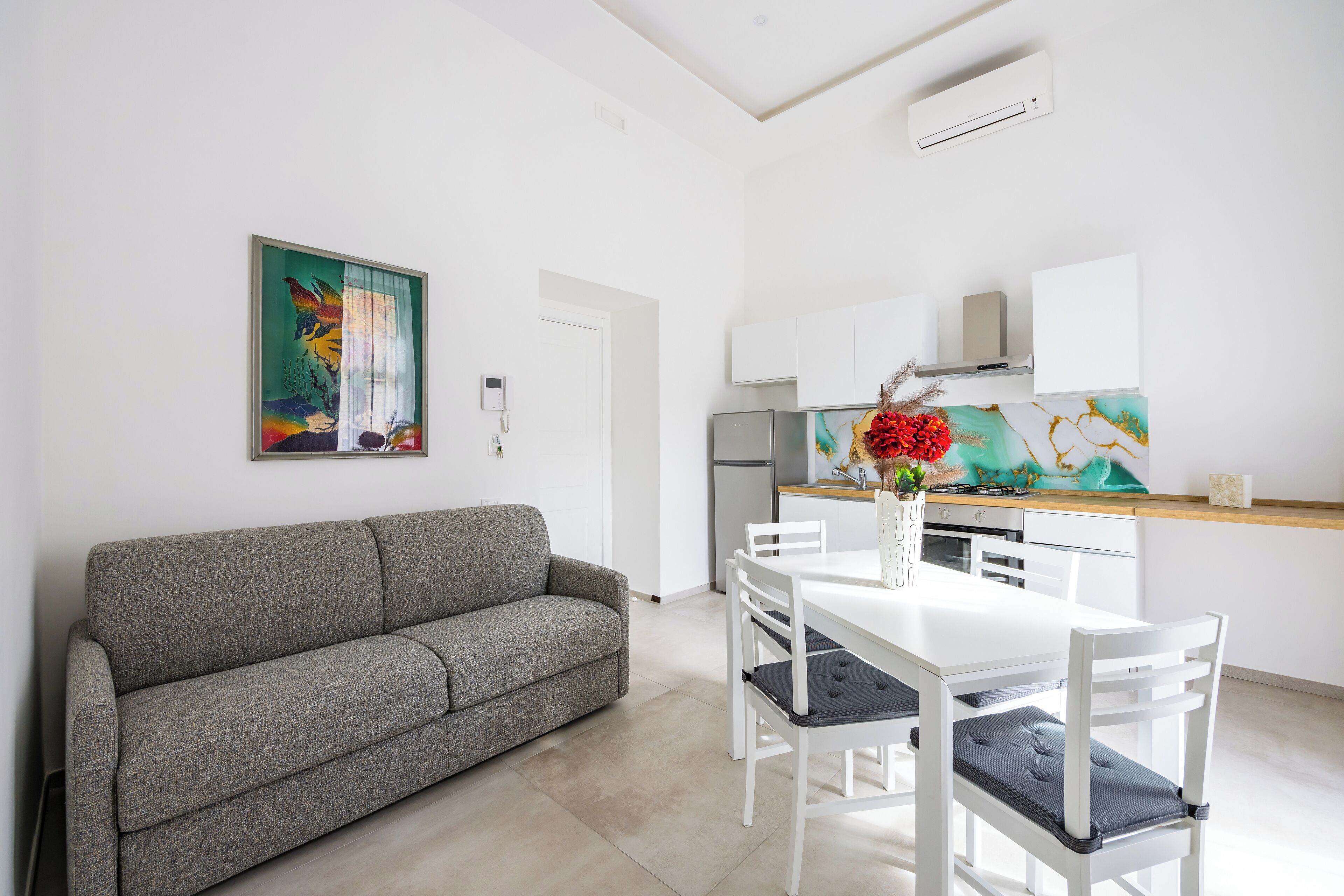 Un salon moderne avec un canapé gris, une table à manger blanche avec quatre chaises, et un coin cuisine doté de meubles blancs et d’un crédence colorée.