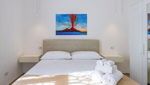 A modern bedroom with a large bed, white linens, a headboard, bedside tables, and a painting of a volcano. - Maison Emanuele Double Apartment (Naples)