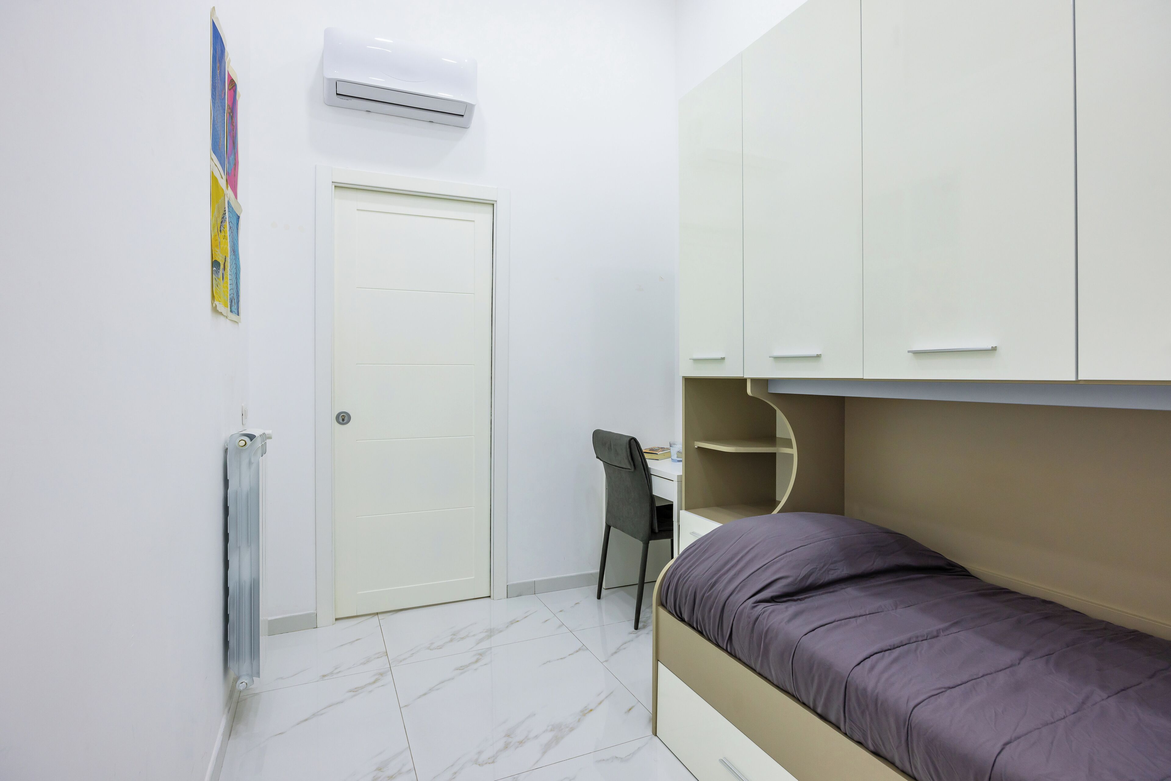 A bedroom with a bed, a desk, and a chair. There is a wardrobe with multiple drawers and a radiator on the wall.