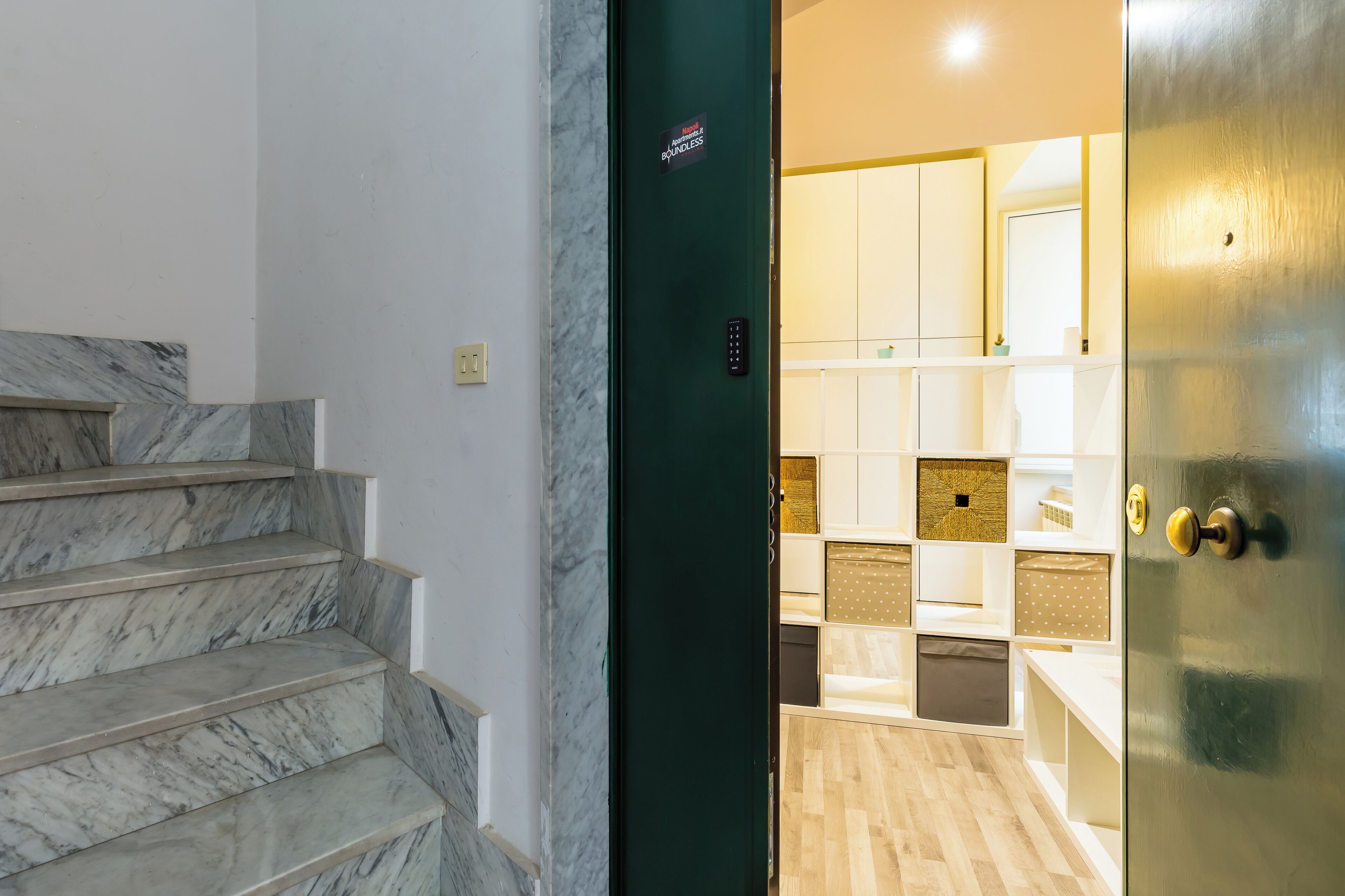 A staircase with marble steps leading to a room with a green door, a control panel, and a kitchen area with white cabinets and wooden flooring.