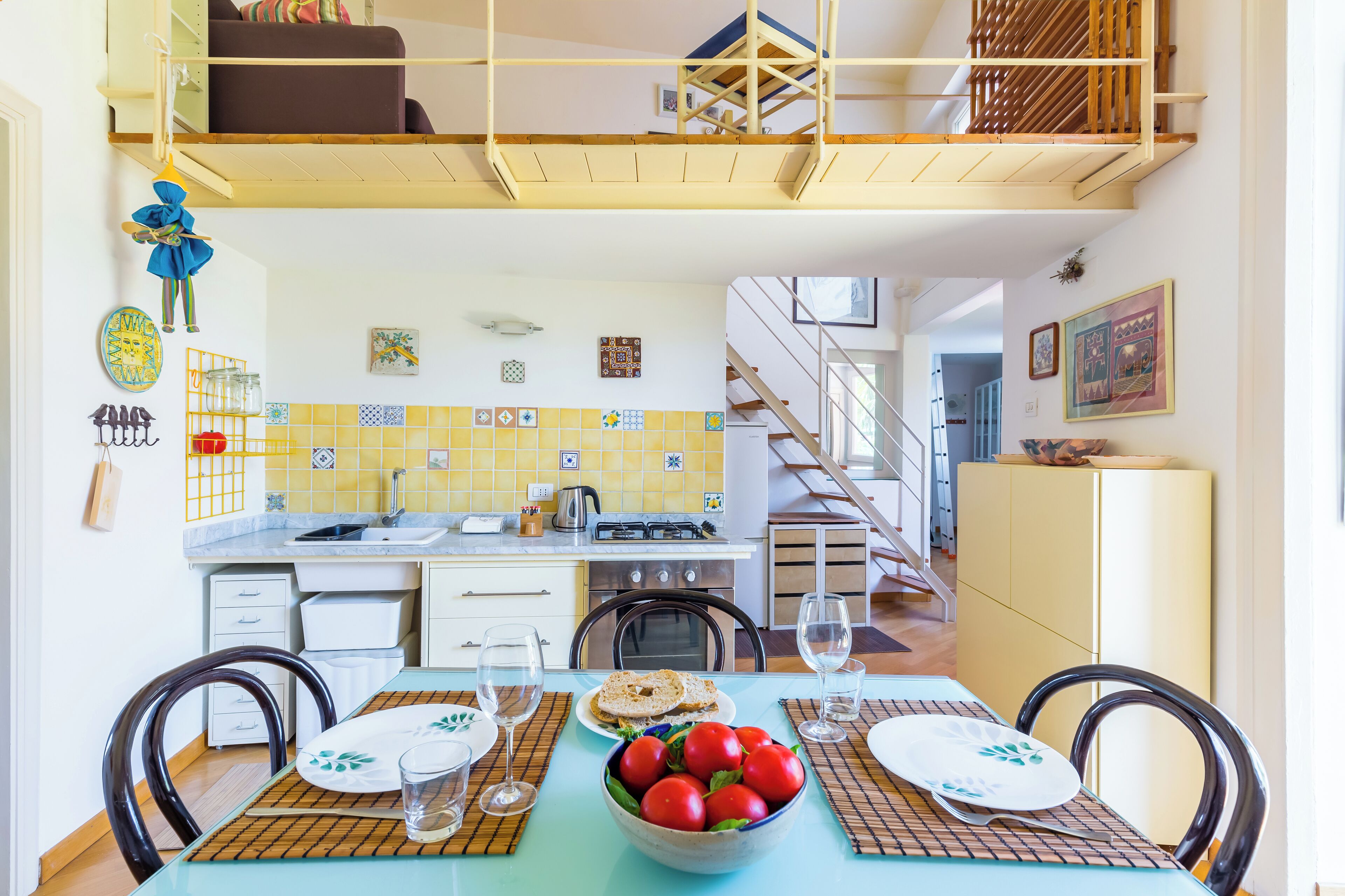 A dining area with a table set for four, a kitchen in the background, and a staircase leading to an upper level.