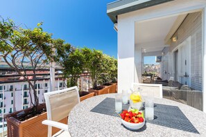 A round table with a bowl of fruit and a pitcher of milk, surrounded by chairs, on a balcony with a city view.