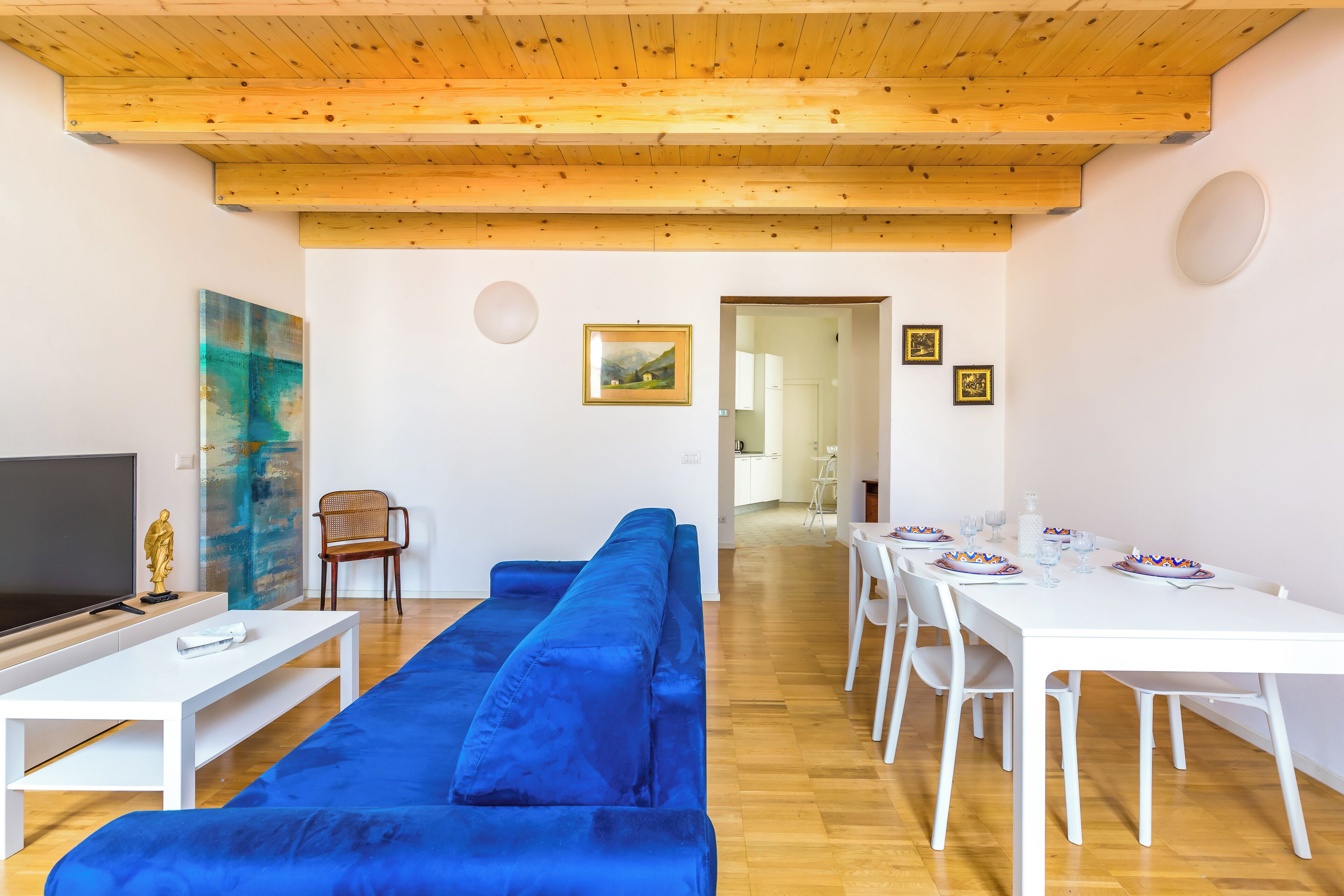 A modern living room with a blue sofa, white dining table, and wooden ceiling beams.