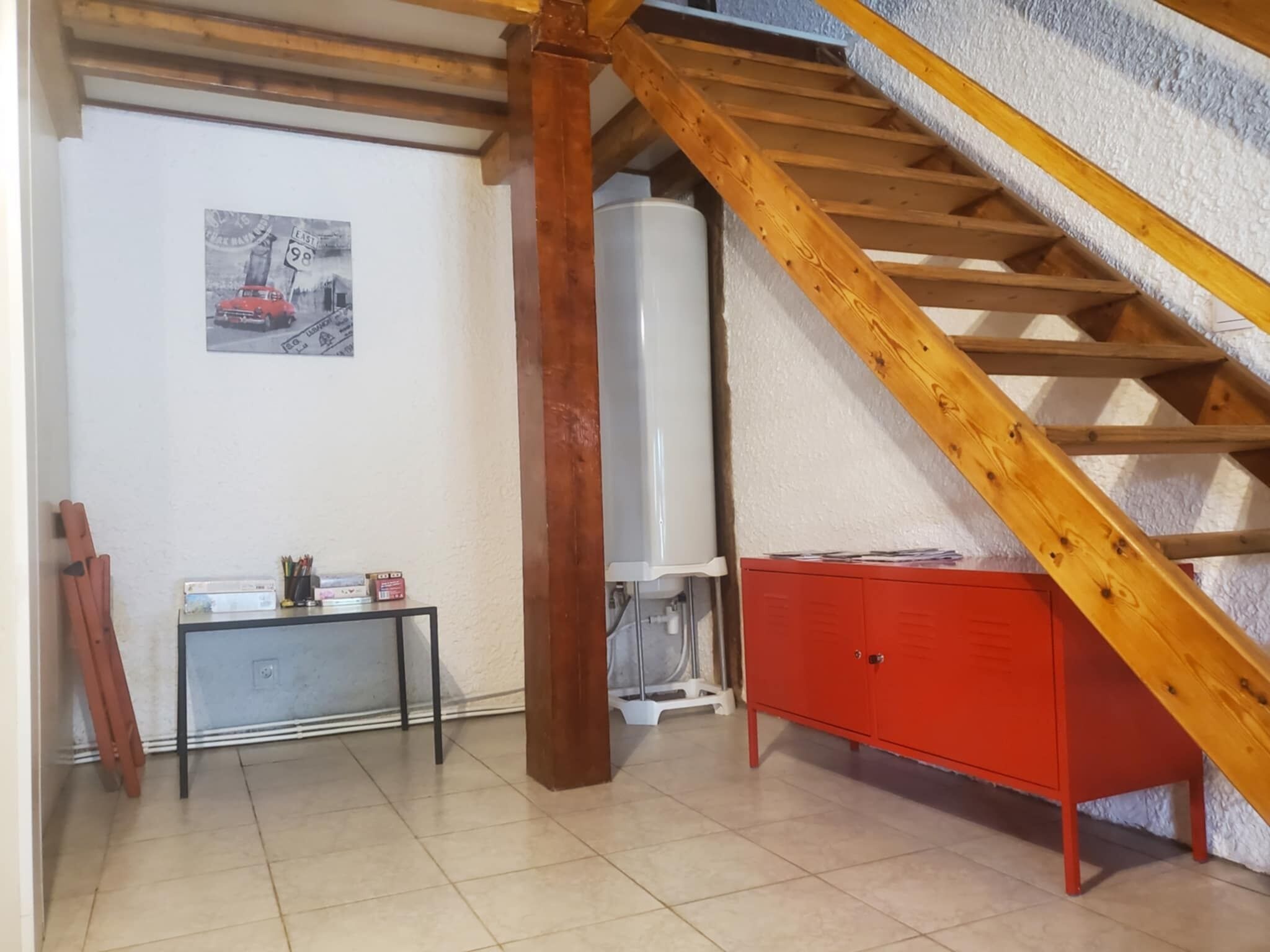 Une pièce avec un escalier en bois, un meuble rouge, un radiateur blanc et une petite table sur laquelle se trouvent divers objets.