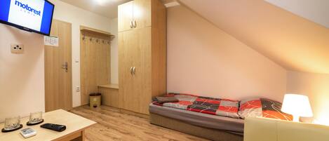 A small, well-lit room with a bed, a desk with a computer and remote controls, a wooden wardrobe, and a television displaying 'motorrest LUMIA'.