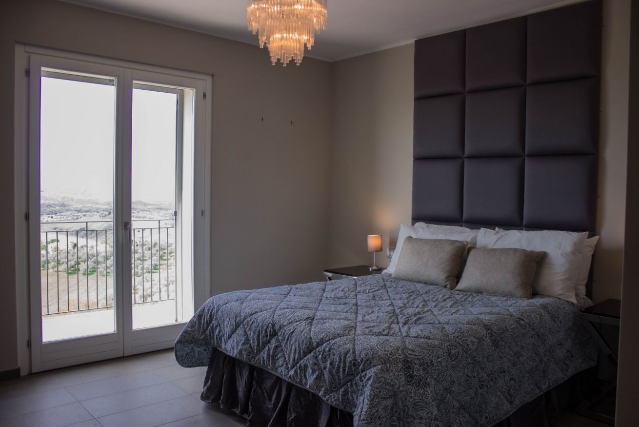 Une chambre à coucher avec un lit, une table de chevet, un lustre et une vue sur l’extérieur, accessible par une porte coulissante.