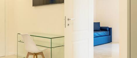 Quarto moderno com escrivaninha de vidro, cadeira branca, sofá azul e um espelho grande na parede.