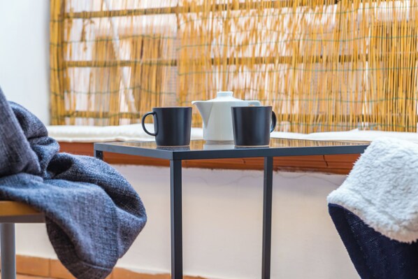 Una mesa con dos tazas negras y una tetera blanca, una manta azul sobre una silla, y una cortina de bambú al fondo.
