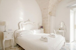 A white, elegantly furnished bedroom with a large bed, a bedside table with a lamp, a nightstand with books, and a mirror on a small table. - Marchesa – Two-Bedroom Suite with Panoramic Terrace in the Historic Center (Monopoli)