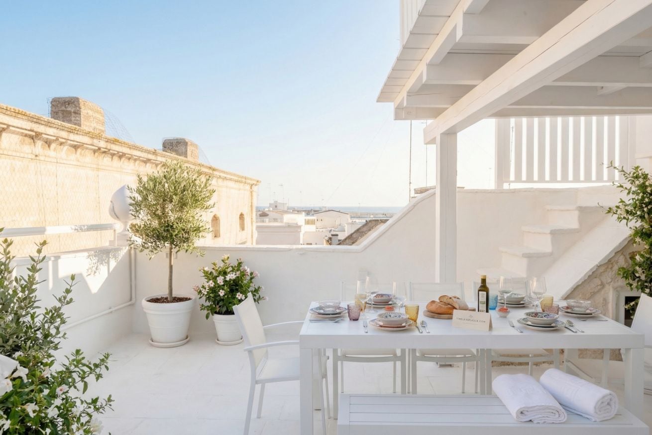A rooftop terrace with a dining table set for two, surrounded by potted plants and white furniture.