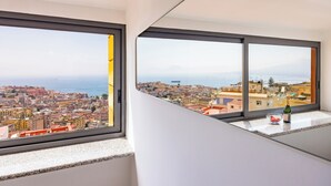 A room with a large window overlooking a coastal cityscape, a small table with a bottle and a bowl of fruit. - Stairway to the Vista Flat (Naples)