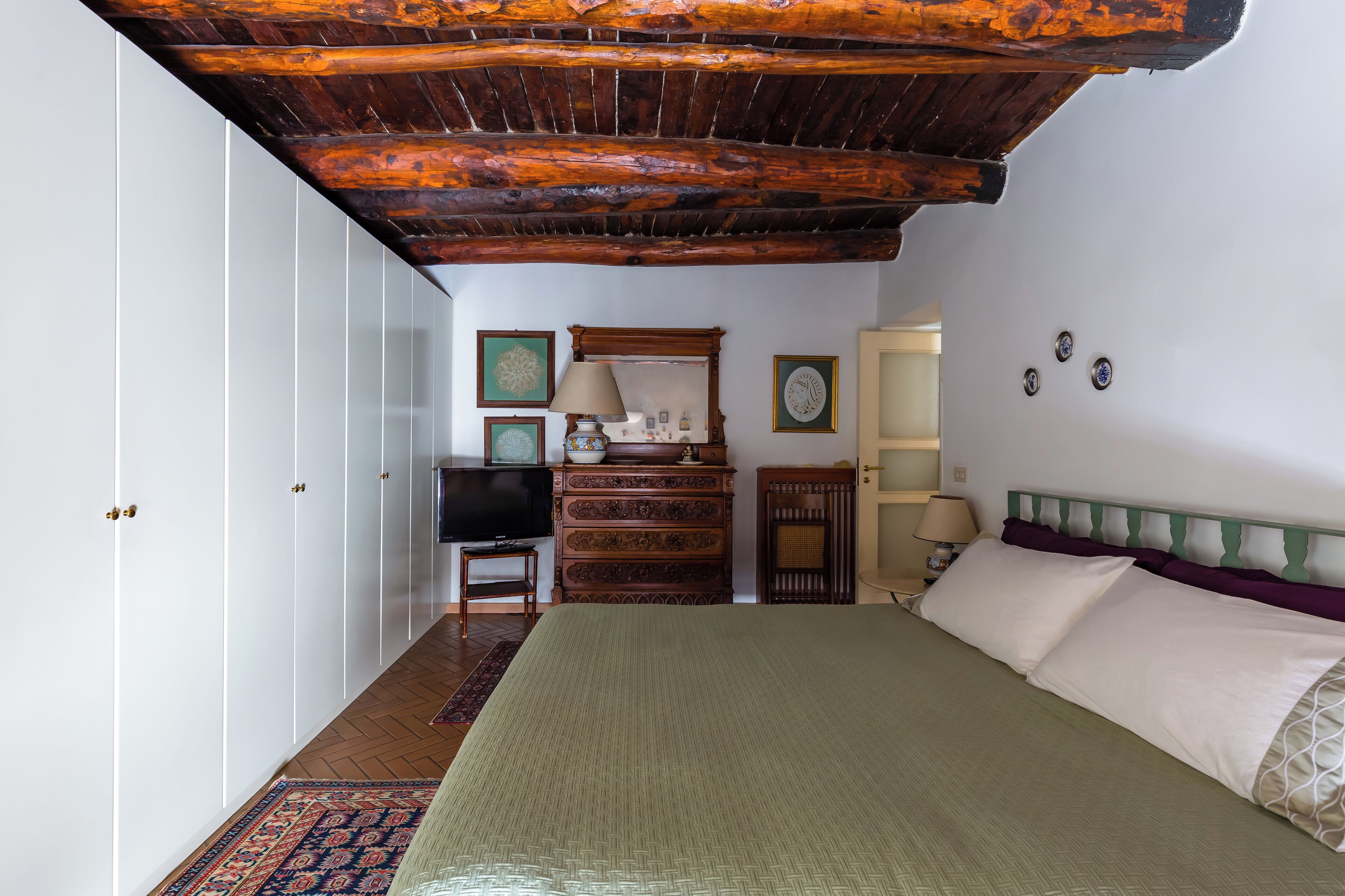 A bedroom with a large bed, wooden ceiling beams, a dresser, a television, and a mirror.