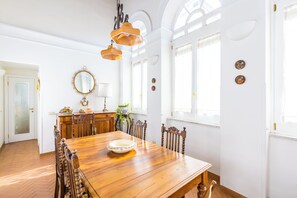 En spisestue med et træbord, stole og en lysekrone. Der er vinduer med gardiner og en dør med frostet glas.