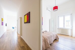 A modern hotel room with a wooden floor, a bed with a beige duvet, a red pendant light, and a large window providing natural light. - Sunny Panoramic Apartment by Napoliapartments (Naples)