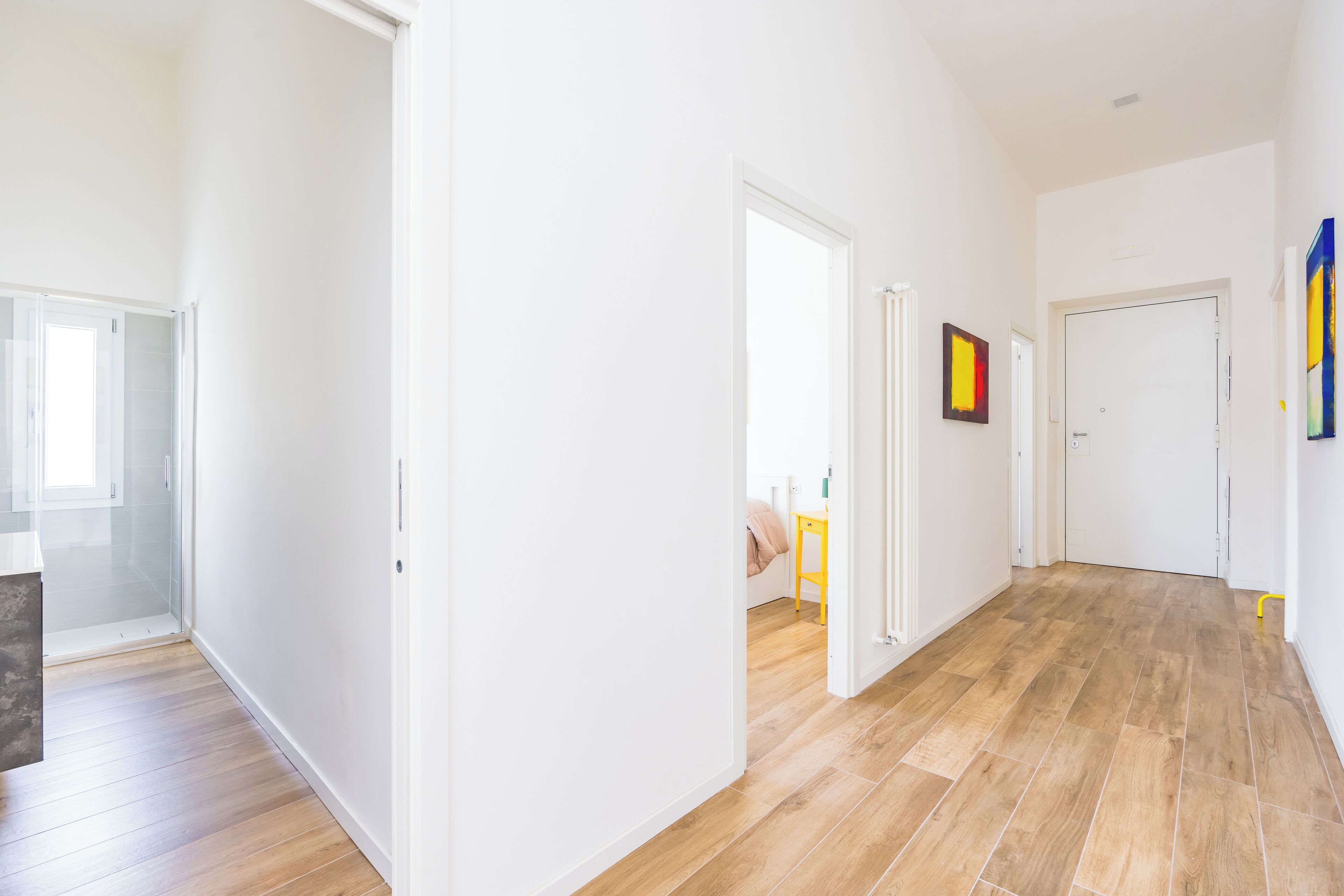 A spacious hallway with wooden flooring and white walls, leading to a bedroom with a bed and a nightstand.
