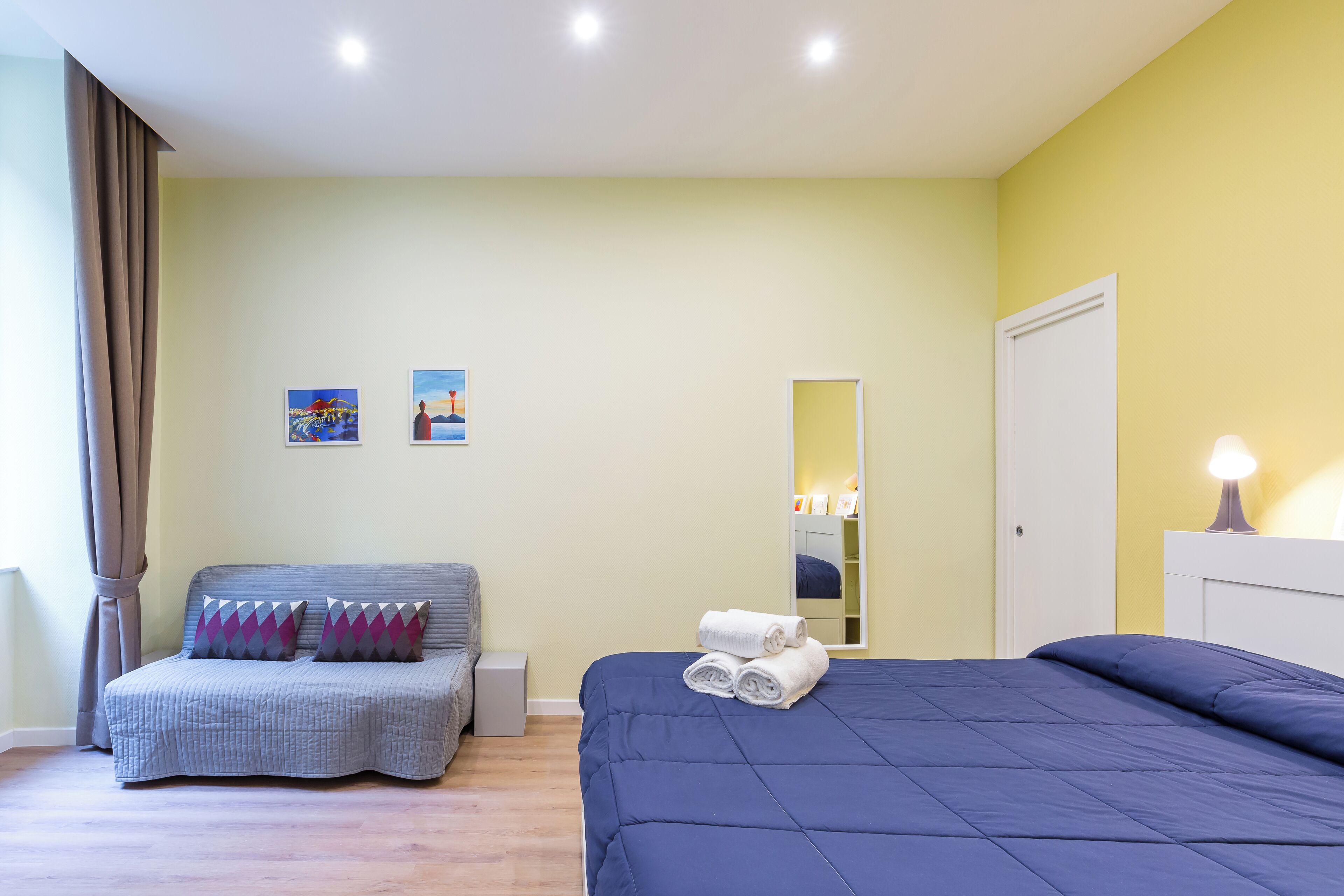 A bedroom with a bed, a sofa, a mirror, and two paintings on the wall.